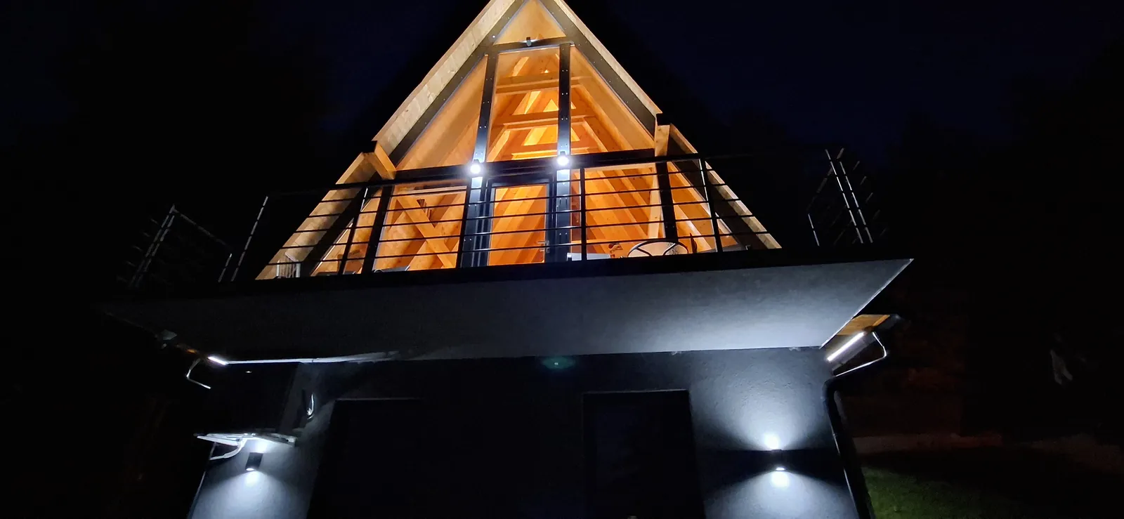 A-frame vikendica noću sa toplo osvetljenim staklenim zabatom.