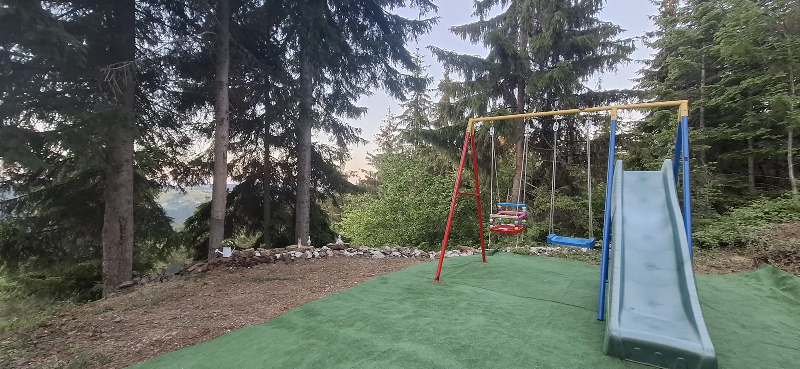 Dečje igralište sa ljuljaškom i toboganom u dvorištu okruženom borovima.